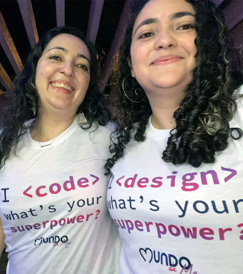 Foto da Mariana e Mariele abraçadas e sorrindo. Ambas com uma camiseta escrito "I code" e "I design" - qual o seu super poder?
